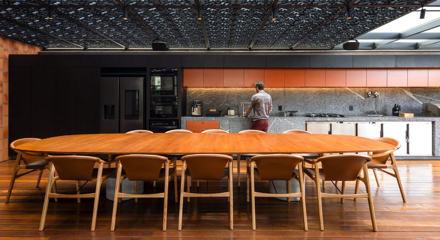 Mesa longa de madeira com cadeiras e a cozinha de armários laranja ao fundo